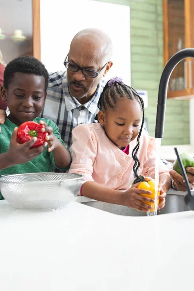 Happy african american grandfather and grandchildren washing vegetables in kitchen. Food, cooking, home, family, togetherness, domestic life and lifestyle, unaltered.
