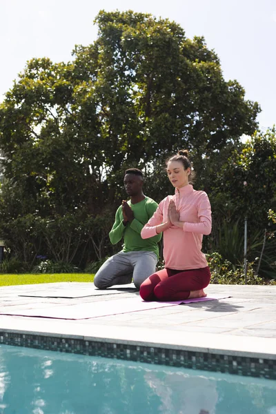 Diverse couple practicing yoga and meditating next to swimming pool in sunny garden, copy space. Lifestyle, togetherness, relationship, relaxation and domestic life, unaltered.