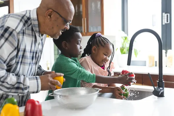 Happy african american grandfather and grandchildren washing vegetables in kitchen, slow motion. Food, cooking, home, family, togetherness, domestic life and lifestyle, unaltered.