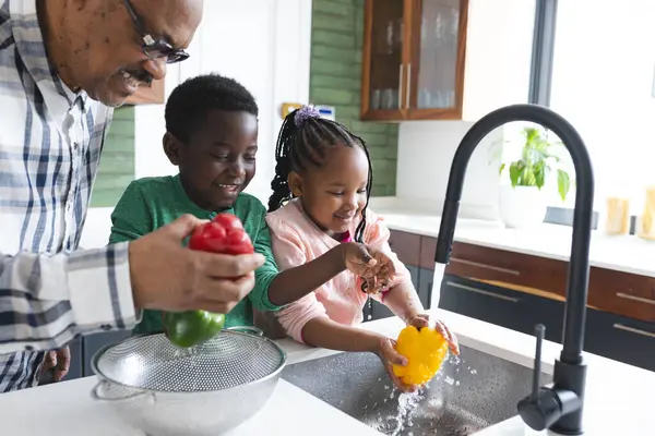 Happy african american grandfather and grandchildren washing vegetables in kitchen, slow motion. Food, cooking, home, family, togetherness, domestic life and lifestyle, unaltered.