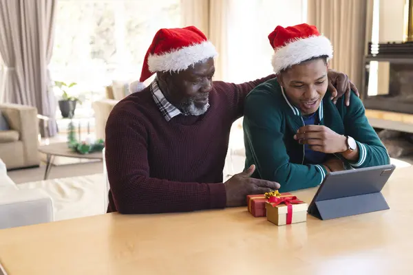 Happy african american father and son in christmas hats having tablet video call at home. Communication, family, friendship, christmas, celebration, togetherness, tradition and lifestyle, unaltered.