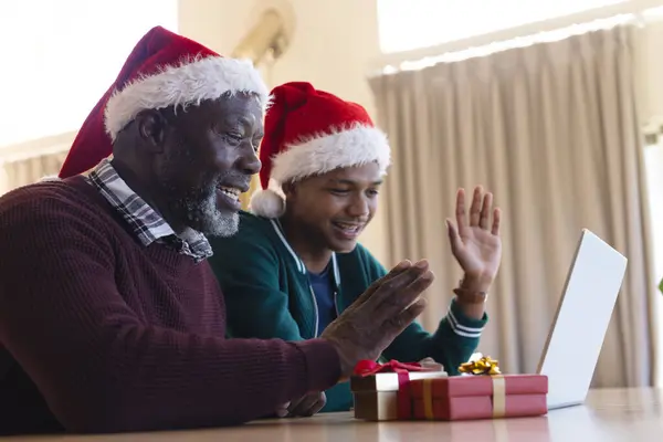 Happy african american father and son in christmas hats having laptop video call at home. Communication, family, friendship, christmas, celebration, togetherness, tradition and lifestyle, unaltered.