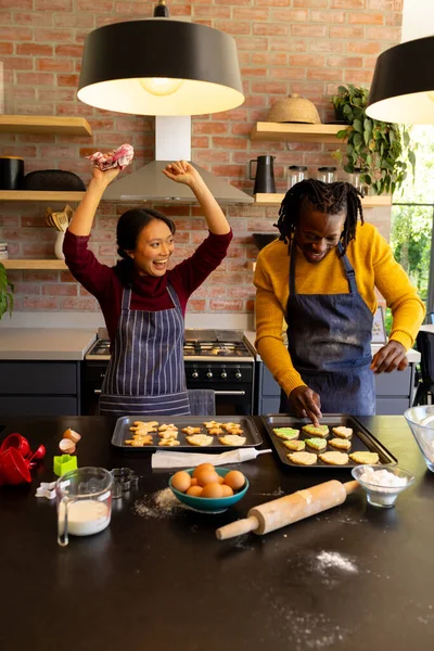 Happy diverse couple in aprons decorating christmas cookies and cheering in kitchen. Fun, cooking, baking, christmas, celebration, togetherness, domestic life and lifestyle, unaltered.