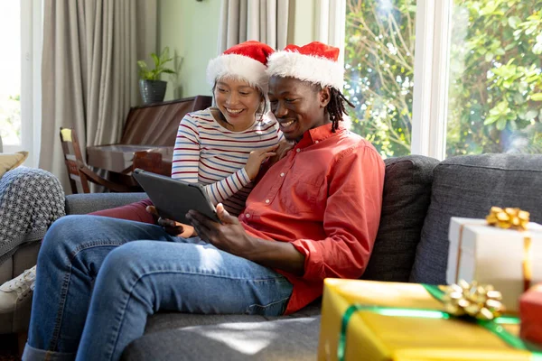 Happy diverse couple in christmas hats having tablet video call smiling in sunny living room. Communication, togetherness, relationship, christmas, celebration, tradition and lifestyle, unaltered.