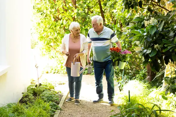 Happy caucasian senior couple walking with flowers and watering can in sunny garden. Retirement, togetherness, domestic life and senior lifestyle, nature, gardening, unaltered.