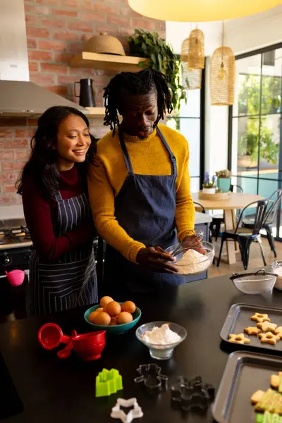 Happy diverse couple in aprons baking christmas cookies in kitchen, copy space. Cooking, baking, food, christmas, celebration, tradition, togetherness, domestic life and lifestyle, unaltered.