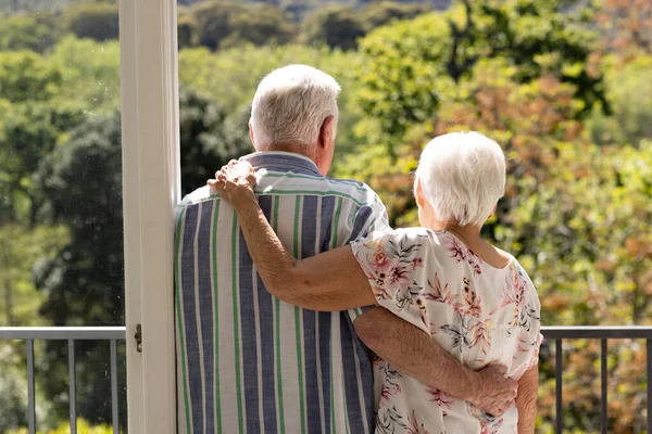Back view of caucasian senior couple embracing and looking ahead on balcony on sunny day. Retirement, togetherness, wellbeing, domestic life and senior lifestyle, unaltered.