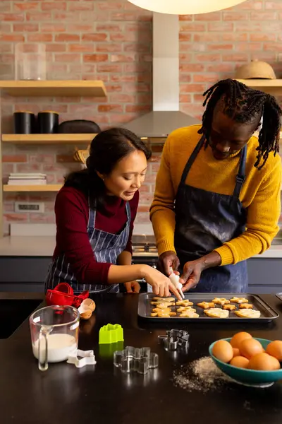 Happy diverse couple in aprons decorating christmas cookies in kitchen, copy space. Cooking, baking, food, christmas, celebration, tradition, togetherness, domestic life and lifestyle, unaltered.