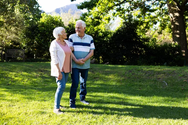 Happy caucasian senior couple walking, embracing and discussing in sunny garden, copy space. Retirement, togetherness, domestic life and senior lifestyle, nature, unaltered.
