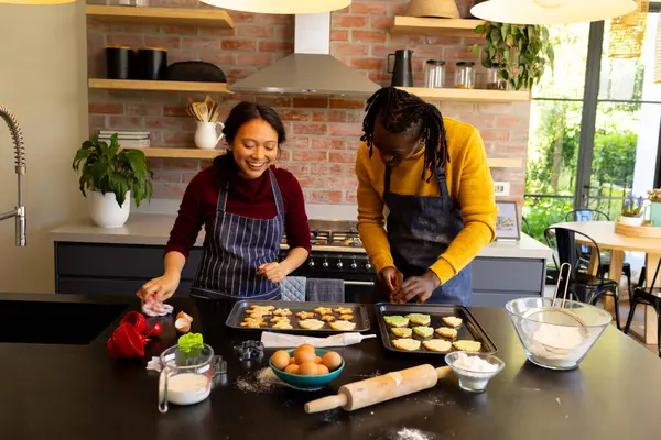 Happy diverse couple in aprons preparing christmas cookies in sunny kitchen. Cooking, baking, christmas, celebration, tradition, togetherness, domestic life and lifestyle, unaltered.