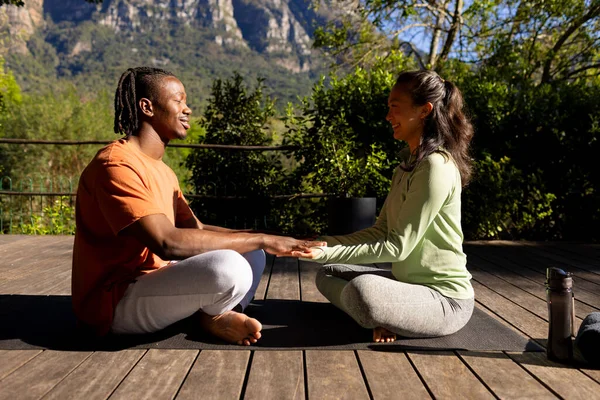 Happy diverse couple practicing yoga meditation sitting holding hands on deck in sunny garden. Yoga, relationship, meditation, togetherness, relaxation, nature and heathy lifestyle, unaltered.