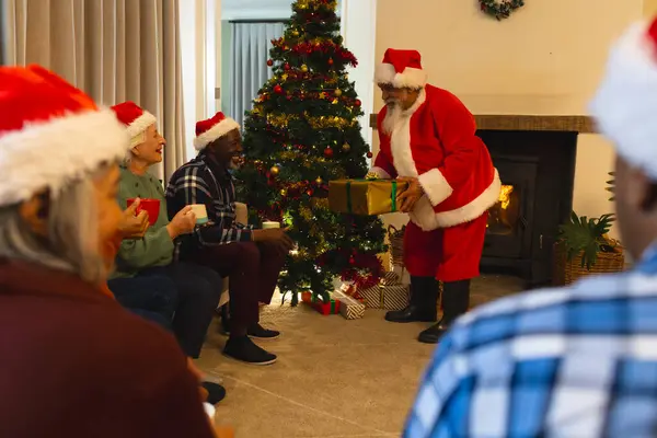 Happy biracial man in santa clothes giving gifts to friends next to christmas tree at home. Retirement, christmas, celebration, friendship, togetherness and senior lifestyle, unaltered.