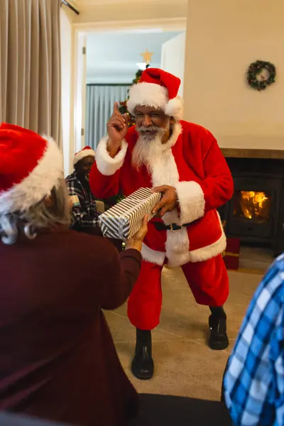 Happy biracial man in santa clothes giving gift to friend next to christmas tree at home. Retirement, christmas, celebration, friendship, togetherness and senior lifestyle, unaltered.