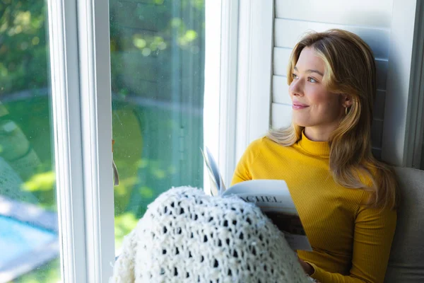 Happy caucasian woman sitting on couch with book and looking out window in sunny room at home. Relaxation, wellbeing, free time and domestic life, unaltered.