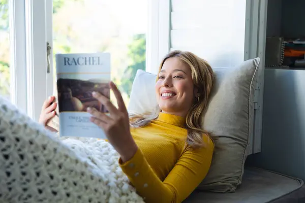 Happy caucasian woman lying on couch, reading book and smiling in sunny room at home. Relaxation, wellbeing, free time and domestic life, unaltered.