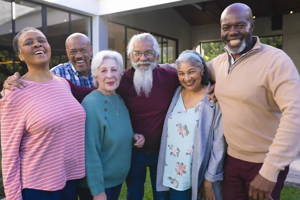 Happy diverse group of senior friends embracing next to house in sunny garden. Retirement, friendship, wellbeing, togetherness and senior lifestyle, unaltered.