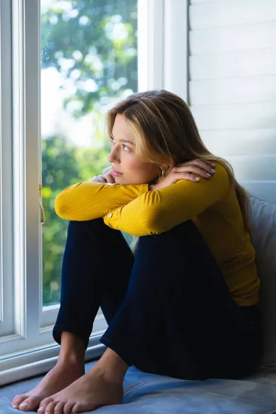 Sad caucasian woman sitting on windowsill and looking out window in sunny room at home. Feelings and emotions, free time and domestic life, unaltered.