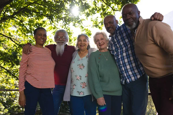 Happy diverse group of senior friends embracing and smiling in sunny garden. Retirement, friendship, wellbeing, nature, togetherness and senior lifestyle, unaltered.