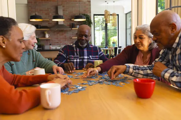 Happy diverse group of senior friends playing with jigsaw puzzles in sunny dining room at home. Retirement, friendship, wellbeing, activities, togetherness and senior lifestyle, unaltered.