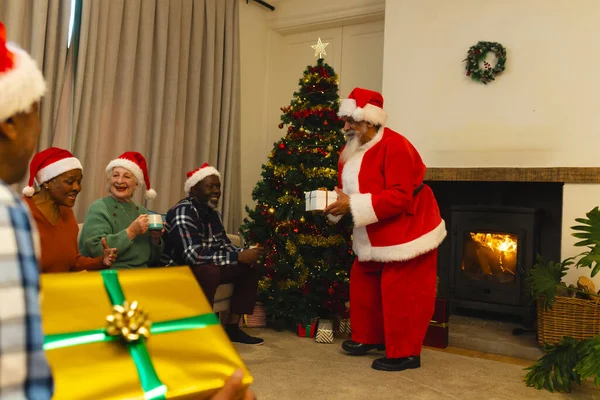 Happy biracial man in santa clothes giving gifts to friends next to christmas tree at home. Retirement, christmas, celebration, friendship, togetherness and senior lifestyle, unaltered.
