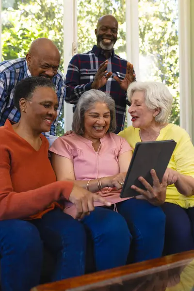 Happy diverse group of senior friends having video call and laughing in sunny living room. Retirement, friendship, domestic life and senior lifestyle, communication, technology, unaltered.