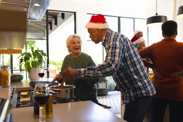 Happy diverse friends couple in santa hat preparing food in sunny kitchen at home. Retirement, friendship, christmas, celebration cooking, eat, senior lifestyle, unaltered.
