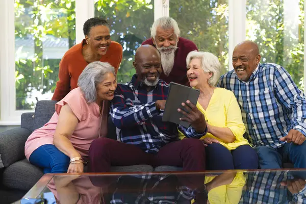Happy diverse group of senior friends using tablet and laughing in sunny living room. Retirement, friendship, domestic life and senior lifestyle, communication, unaltered.
