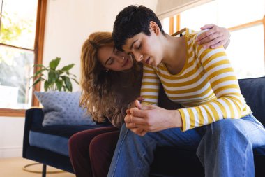 Sad biracial lesbian couple embracing and comforting, sitting on couch in sunny living room. Depression, mental health, support, care, gay, relationship, togetherness and domestic life, unaltered. clipart