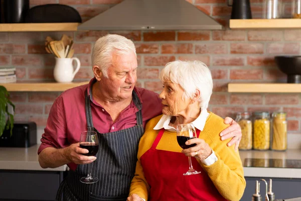 Happy caucasian senior couple cooking, drinking wine and embracing in kitchen. Drinking, togetherness, retirement, cooking, domestic life and senior lifestyle, unaltered.