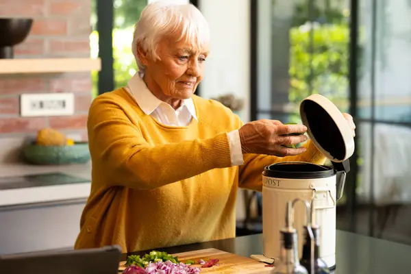 Happy caucasian senior woman preparing food, composting vegetable waste in kitchen. Ecology, recycling, retirement, cooking, healthy eating, domestic life and senior lifestyle, unaltered.