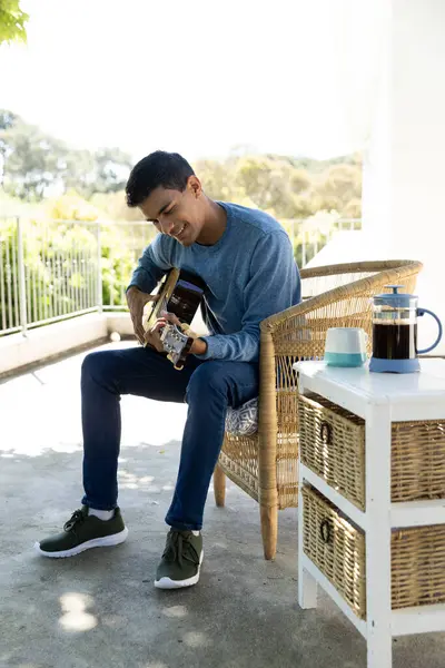 Happy biracial man sitting on balcony playing guitar at home. Hobbies, music, lifestyle and domestic life, unaltered.