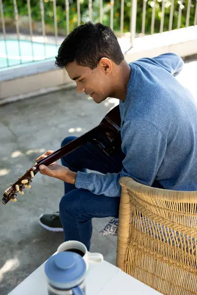 Happy biracial man sitting on balcony playing guitar at home. Hobbies, music, lifestyle and domestic life, unaltered.