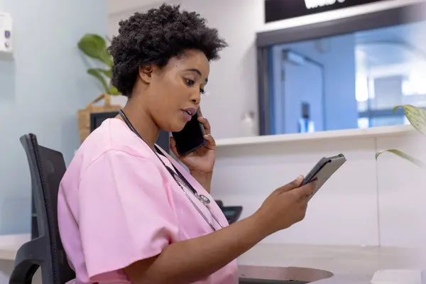 非洲裔美国女医生在医院接待处用智能手机和平板电脑交谈 通信和医疗服务 未加改动 — 图库照片