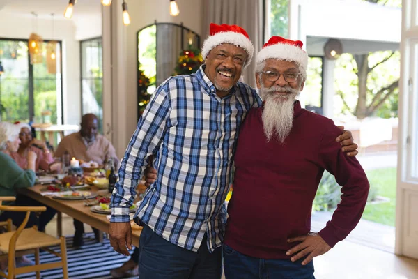 Happy diverse senior male friends in santa hats embracing at christmas meal with friends. Retirement, christmas, celebration, friendship, togetherness and senior lifestyle, unaltered.
