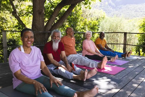 Happy diverse group of senior friends sitting on mats at yoga class in sunny garden, copy space. Retirement, friendship, wellbeing, yoga, fitness and healthy senior lifestyle, unaltered.