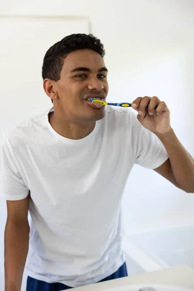 Biracial man brushing teeth looking in mirror in bathroom at home. Self care, hygiene, lifestyle and domestic life, unaltered.