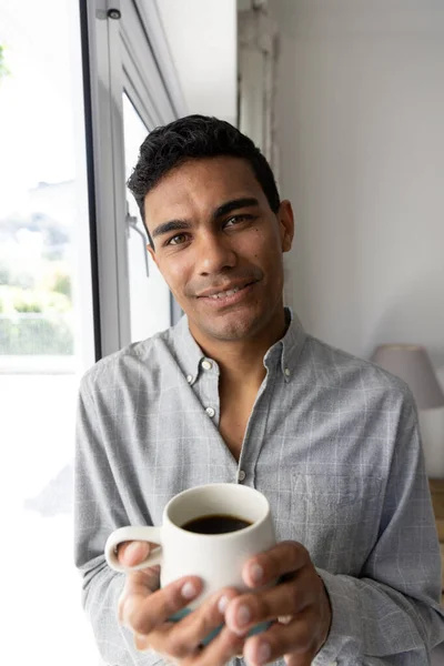 Portrait of happy biracial man holding mug next to window at home. Lifestyle and domestic life, unaltered.