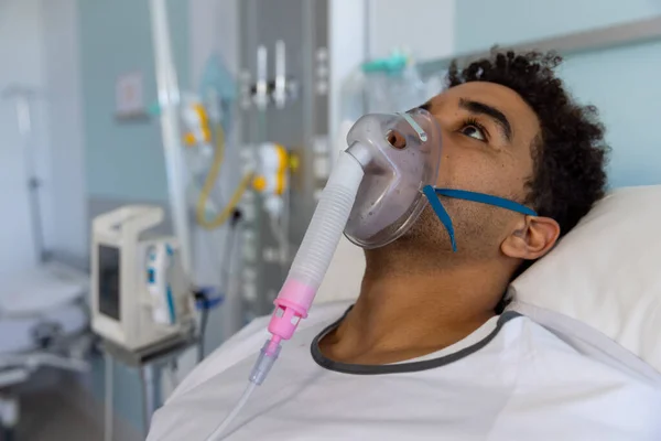 Biracial male patient with oxygen mask lying in bed in sunny hospital room. Medicine, healthcare and medical services, unaltered.