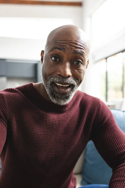 Happy surprised african american mature man having video call in sunny living room at home. Communication, technology, domestic life and lifestyle, unaltered.