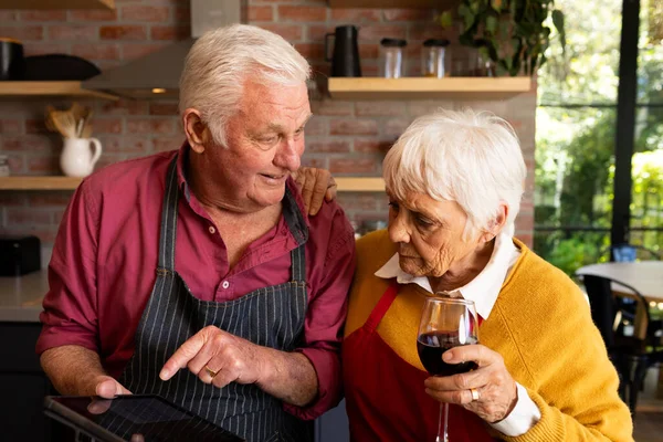 Happy caucasian senior couple cooking, drinking wine and using tablet in kitchen. Communication, recipe, togetherness, retirement, cooking, domestic life and senior lifestyle, unaltered.