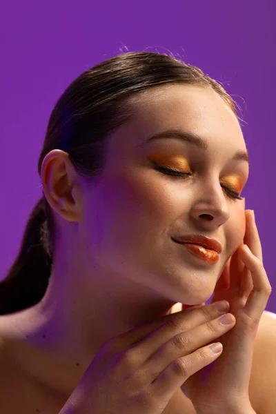 Caucasian woman wearing orange eye shadow and lip gloss on purple background. Cosmetics, makeup, female fashion and beauty, unaltered.