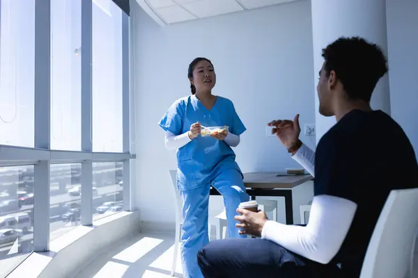 Diverse male and female doctors talking, drinking coffee and eating lunch in sunny hospital. Medicine, healthcare and medical services, unaltered.