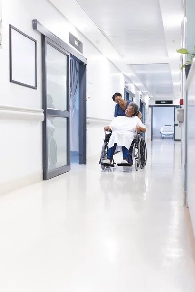 African american female doctor walking with senior female patient in wheelchair in hospital corridor. Medicine, healthcare and medical services, unaltered.
