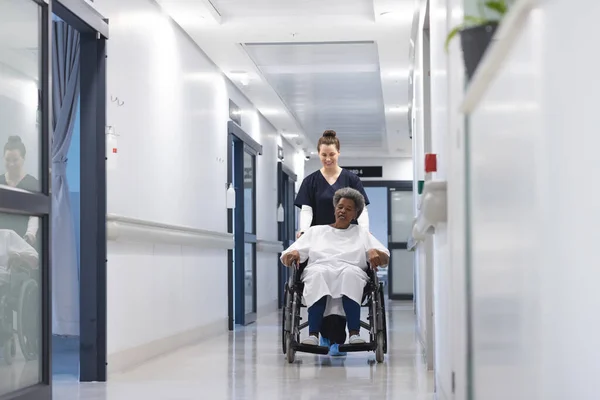 Diverse female doctor walking with senior female patient in wheelchair in hospital corridor. Medicine, healthcare and medical services, unaltered.