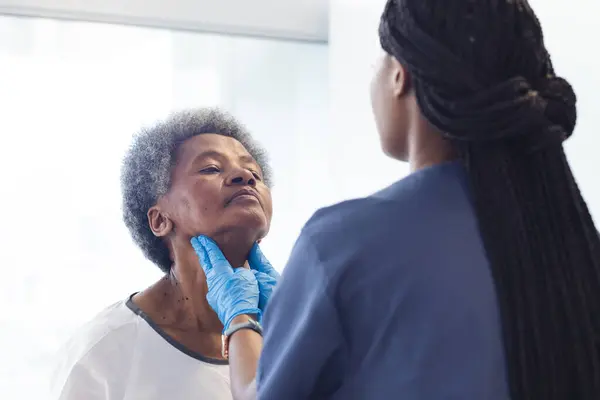 African american female doctor testing neck of senior female patient in hospital room. Medicine, healthcare and medical services, unaltered.