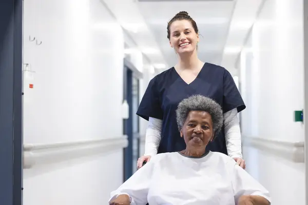 Happy diverse female doctor walking with senior female patient in wheelchair in hospital corridor. Medicine, healthcare and medical services, unaltered.