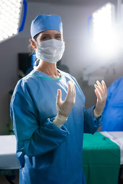 Portrait of caucasian female doctor with face mask and protective gloves in hospital operating room. Medicine, healthcare and medical services, unaltered.
