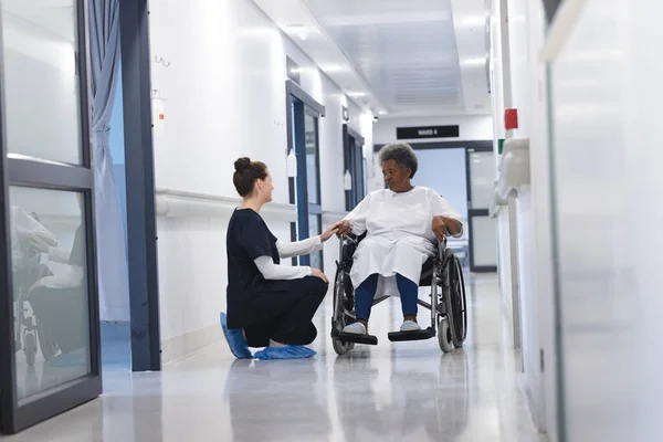 Diverse female doctor talking with senior female patient in wheelchair in hospital corridor. Medicine, healthcare and medical services, unaltered.