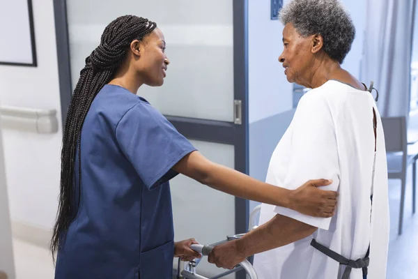 African american female doctor talking with senior female patient with crutches in hospital corridor. Medicine, healthcare and medical services, unaltered.