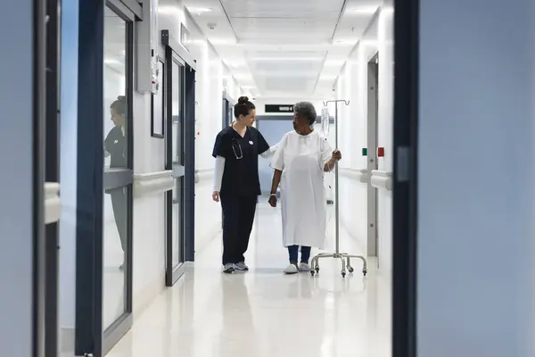 Diverse female doctor talking with senior female patient with drip in hospital corridor. Medicine, healthcare and medical services, unaltered.